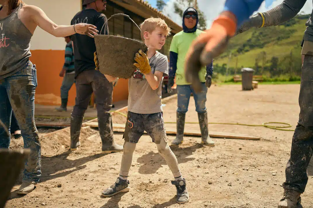 A Young Boy Participates In A Community Service Project, Carrying A Bucket With Other Volunteers In The Background.