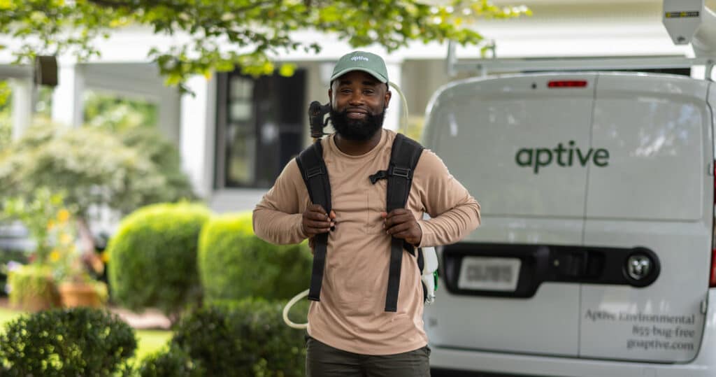 Aptive Pest Control Specialist Wearing A Tan Shirt And Green Aptive Cap, Standing In Front Of An Aptive Van, Smiling And Holding The Straps Of His Backpack Sprayer.