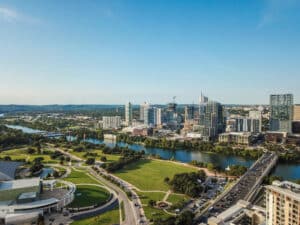 A Detailed Aerial Image Of Downtown Austin, Texas, With The Colorado River Flowing Through The City, Surrounded By Modern Buildings, Green Spaces, And Bridges Under A Clear Blue Sky.