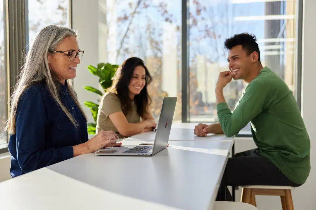 A Diverse Group Of Colleagues At Aptive Environmental Engaging In A Friendly And Productive Discussion Around A Laptop In A Bright Office Space.