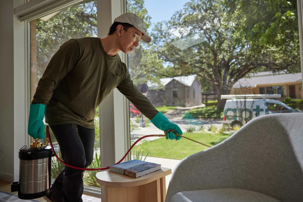 An Aptive Pest Control Specialist, Wearing Protective Gloves And A Cap, Is Spraying Pesticide Indoors Near A Window With A Garden Visible Outside.