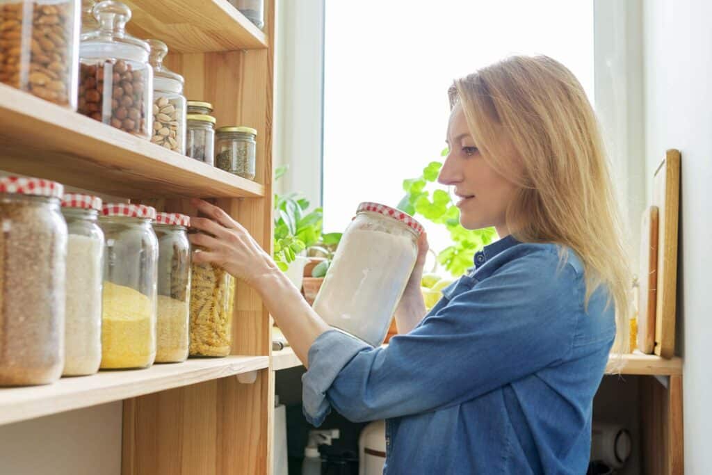 A Woman Organizing A Pantry Shelf Filled With Jars Of Dry Goods Such As Pasta, Flour, And Grains, With Plants In The Background.