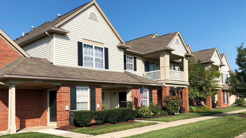 Well-Maintained Townhouse Complex In Farmington Hills, Featuring Red Brick And Beige Siding, Adorned With American Flags, On A Clear Sunny Day.