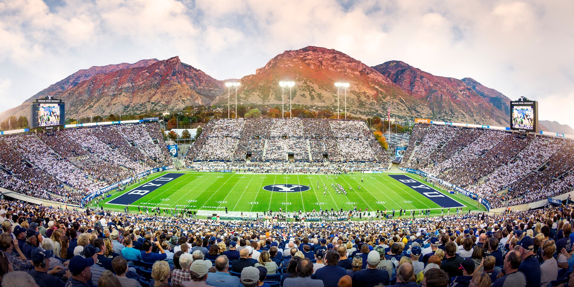 A large crowd fills the stands of a football stadium, with Brigham Young University (BYU) logos on the field. The scene is set against a backdrop of picturesque mountains under a sky with light clouds.