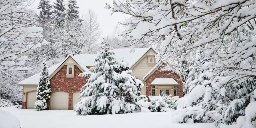 A Picturesque House Covered In Snow, Surrounded By Snow-Laden Trees And Shrubs, Creating A Serene Winter Wonderland Scene.