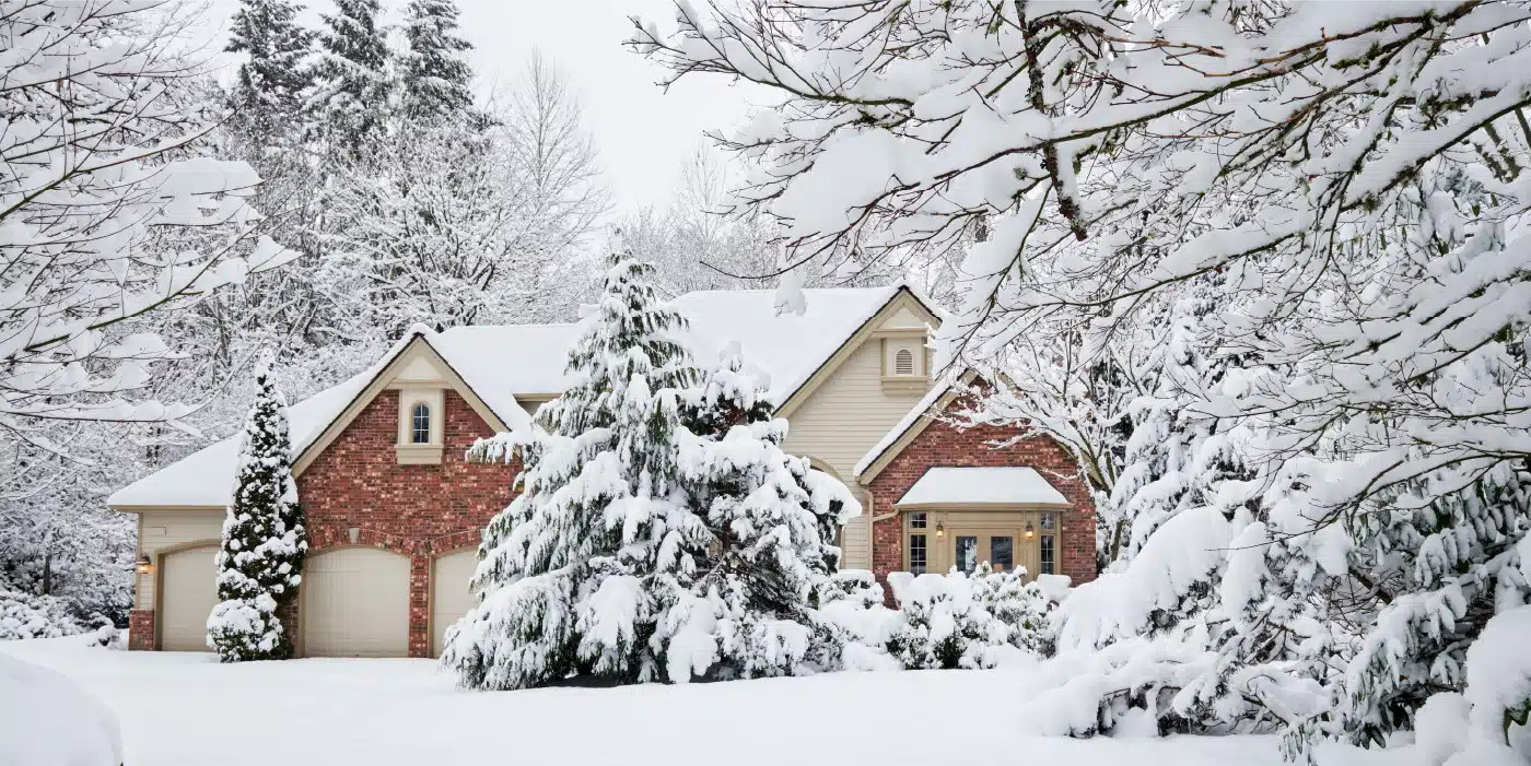 A Snow-Covered House Surrounded By Trees In A Winter Landscape. The House Has A Brick And Beige Exterior, And The Trees Are Heavily Laden With Snow, Creating A Picturesque Winter Scene.