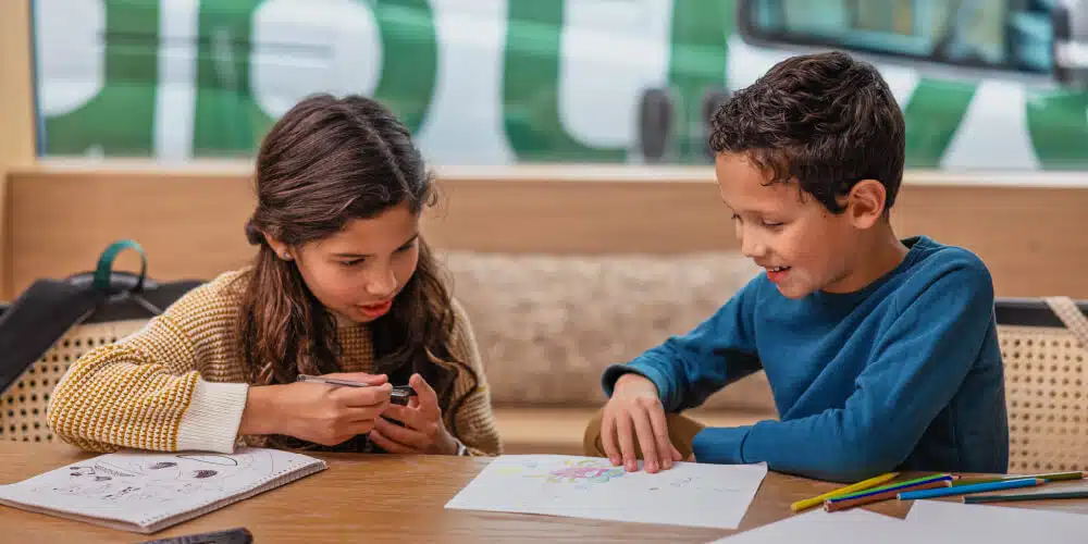 Two Children Sitting At A Table And Engaging In Creative Activities. One Child Is Drawing With Colored Pencils While The Other Is Examining Something With A Magnifying Glass. The Background Shows A Blurred View Of A Green And White Vehicle.