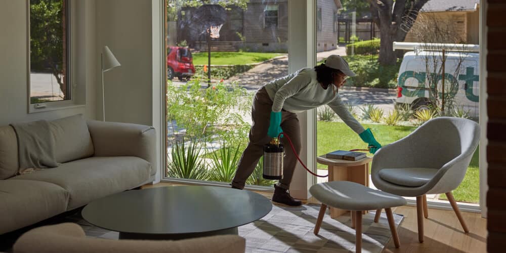 Aptive Pest Control Specialist Treating A Living Room, Spraying Near A Small Table Between A Sofa And A Modern Chair, With A View Of A Sunny Suburban Street Through The Window.