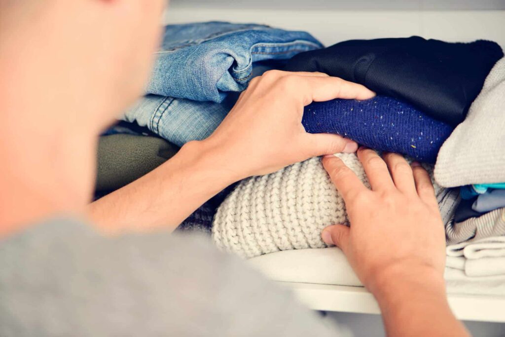 Person Organizing A Stack Of Folded Clothes On A Shelf, Including Items Like Sweaters And Jeans.