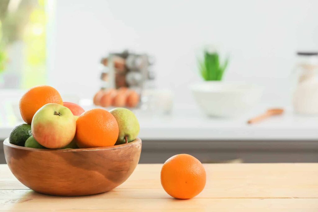 A Wooden Bowl Filled With Apples, Oranges, Pears, And Avocados On A Wooden Table, With An Orange Placed Next To The Bowl. The Background Shows A Modern Kitchen With Blurred Elements.