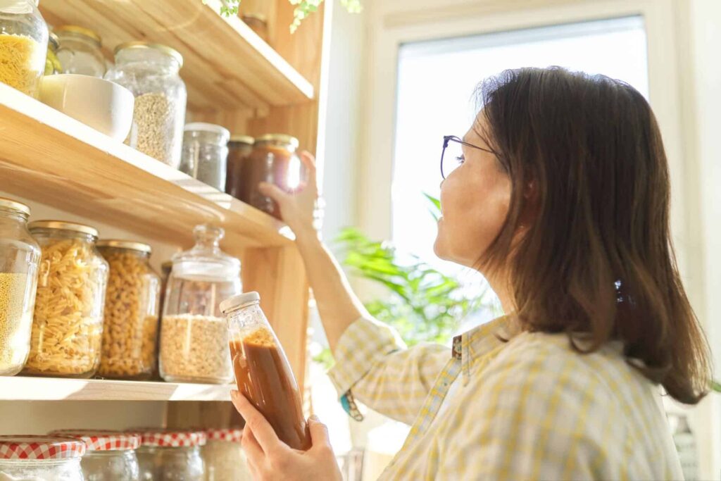 A Woman With Glasses Is Organizing Jars Of Dry Goods And Preserves On Wooden Shelves In A Bright, Sunlit Kitchen.