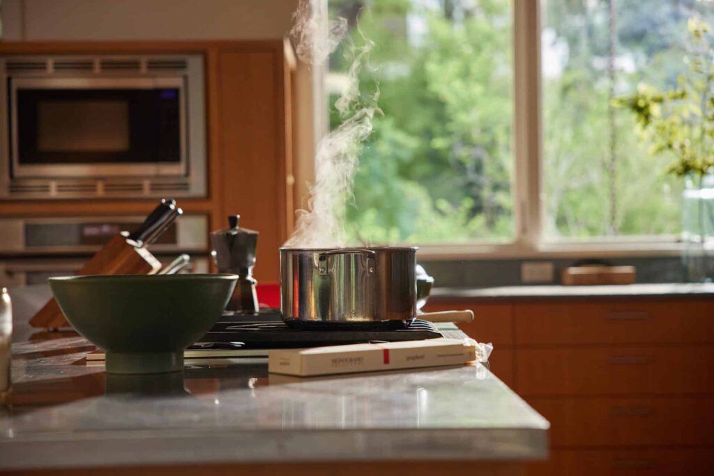 A Modern Kitchen With A Large Window And Wooden Cabinets, Featuring A Stainless Steel Pot On The Stove With Steam Rising From It, A Green Bowl, And A Box Of Pasta On The Countertop.