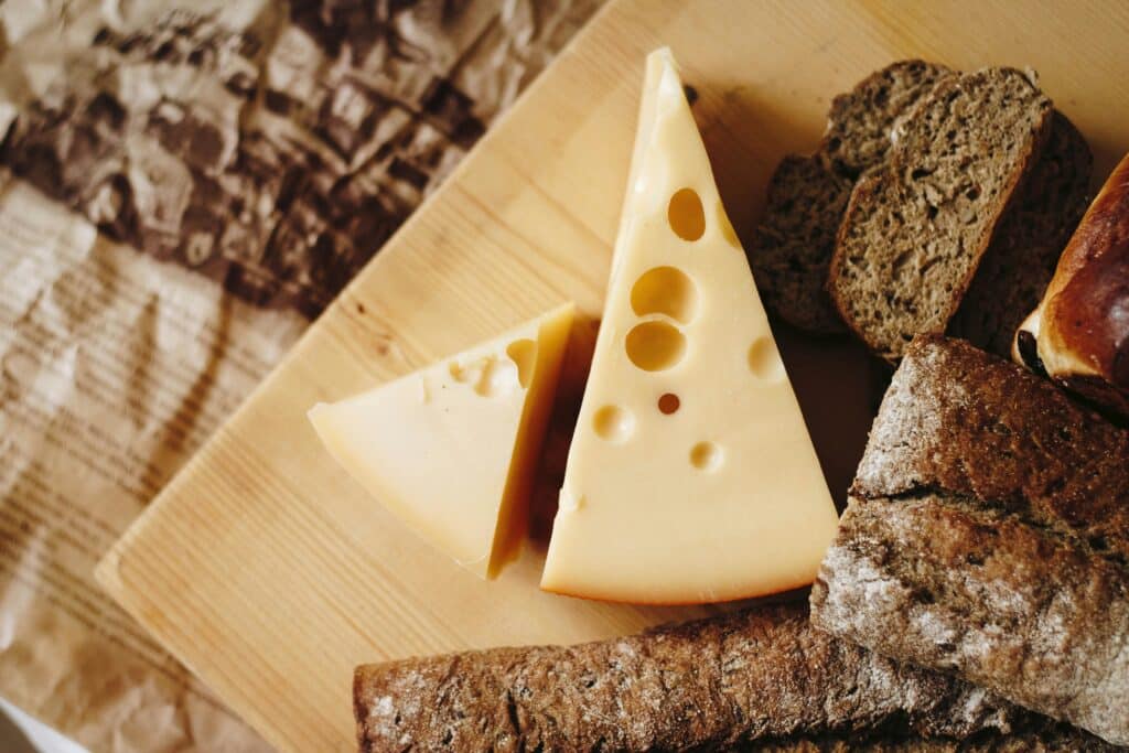 A Close-Up Image Of A Wooden Board With Slices Of Swiss Cheese Featuring Holes, Surrounded By Various Types Of Bread.