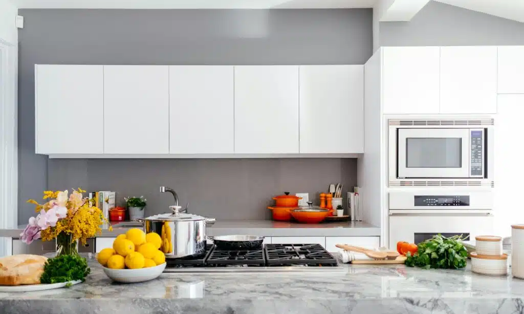 A Modern Kitchen With White Cabinets, A Gray Backsplash, And Stainless Steel Appliances. The Kitchen Features A Gas Stovetop With A Shiny Pot And A Bowl Of Lemons On The Marble Countertop. Brightly Colored Orange Cookware And Various Fresh Ingredients, Including Bread, Herbs, And Flowers, Add Vibrant Accents To The Space.