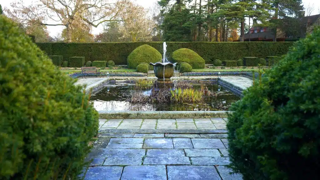 A Well-Maintained Garden Featuring A Central Fountain With Water Flowing From The Top. The Fountain Is Surrounded By Neatly Trimmed Hedges And Bushes, With A Paved Walkway Leading To It. The Background Includes Additional Manicured Hedges, Trees, And A Bench, Creating A Peaceful And Orderly Outdoor Setting.