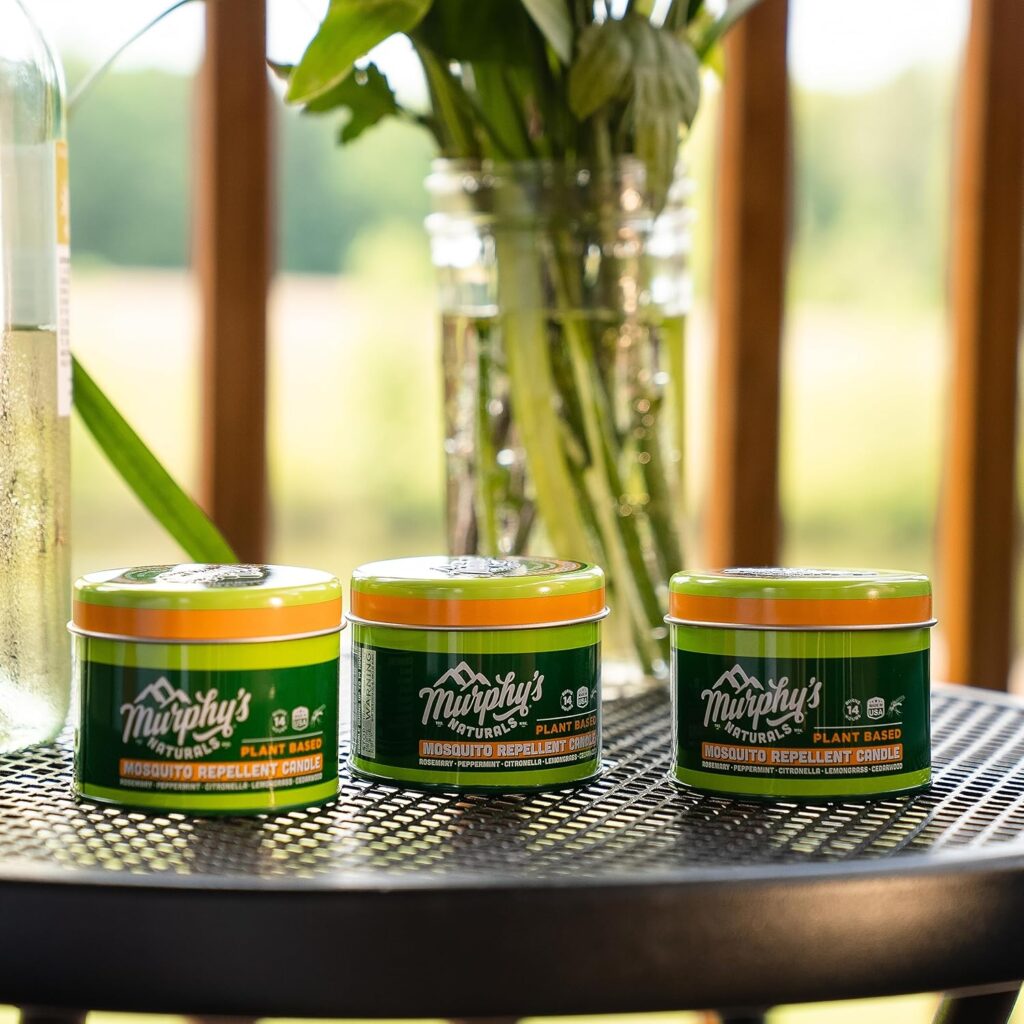 Three Murphy'S Naturals Plant-Based Mosquito Repellent Candles On A Patio Table, With A Vase Of Green Plants In The Background.