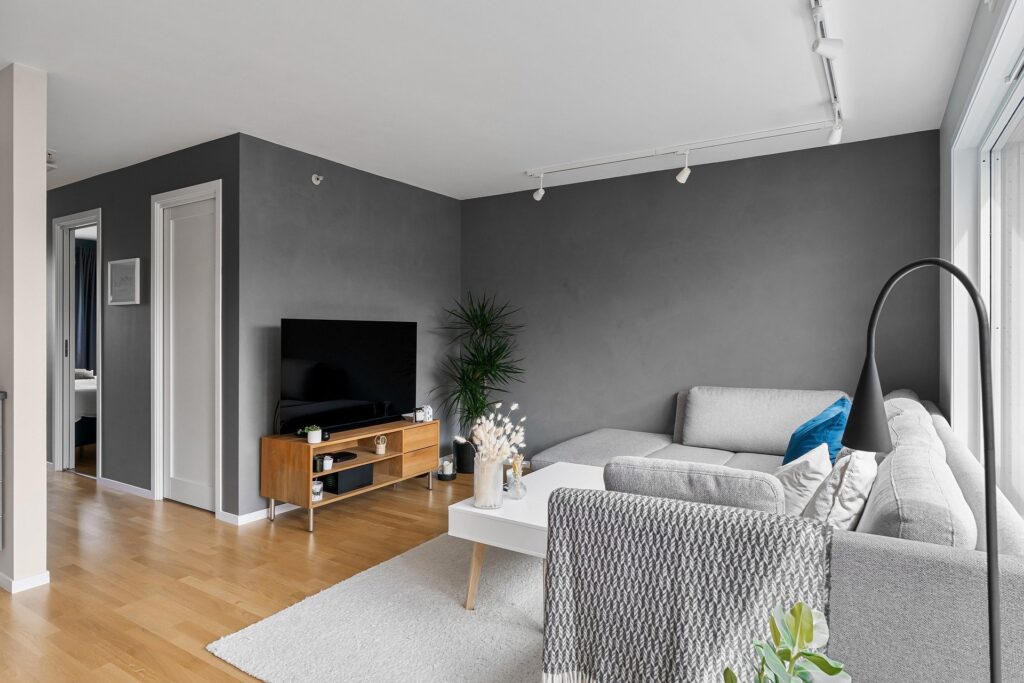 Modern Living Room With Gray Walls, A Light Gray Sectional Sofa, A White Coffee Table, And A Wooden Tv Stand. A Black Flat-Screen Tv And A Potted Plant Are Placed On The Stand.