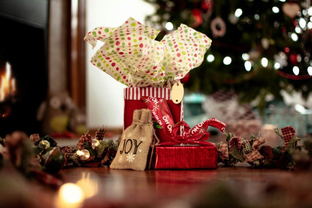 A Festive Red Gift Box With Colorful Polka Dot Tissue Paper And A Burlap Sack Labeled &Quot;Joy,&Quot; Surrounded By Rustic Holiday Decorations, Set Against A Blurred Christmas Tree And Fireplace Background.