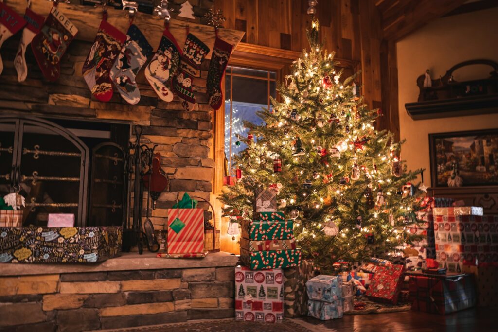 A Cozy Living Room Decorated For Christmas, Featuring A Stone Fireplace Adorned With Stockings And A Brightly Lit Christmas Tree Surrounded By Neatly Wrapped Presents.