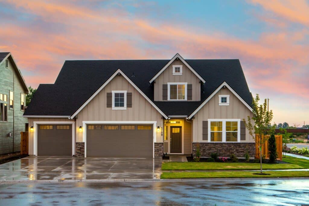 Modern Suburban Home With Beige Siding, Dark Shutters, And A Clean Black Roof, Featuring A Well-Maintained Lawn And A Triple Garage Under A Colorful Evening Sky.