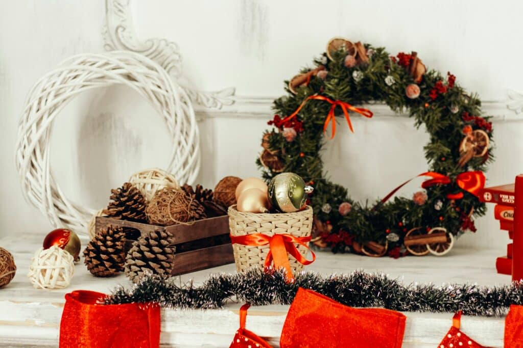 A Holiday-Themed Mantel Decorated With A Festive Wreath, Pinecones, Ornaments, And Red Stockings, With A Rustic Wooden Crate And Wicker Basket Adding To The Seasonal Ambiance.