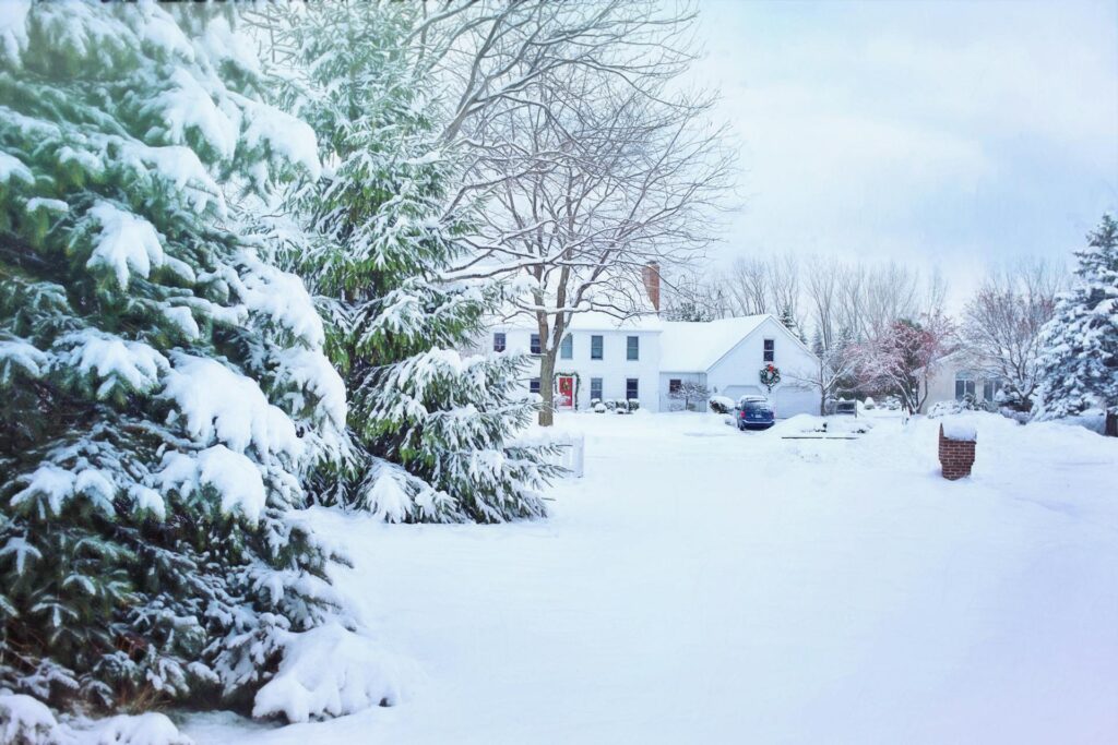 A Snow-Covered Suburban Landscape Featuring A Two-Story White House With A Red Front Door And A Wreath. Snow-Laden Evergreen Trees Frame The Scene, Creating A Picturesque Winter Setting.