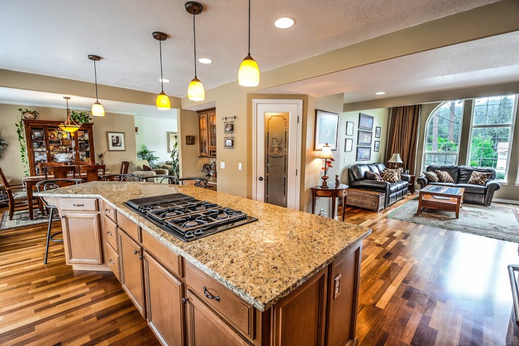 A Detailed Image Of A Modern, Open-Plan Kitchen Featuring Granite Countertops, A Gas Stove, And Warm Pendant Lighting. The Background Shows A Living And Dining Area With Large Windows And Wooden Flooring.