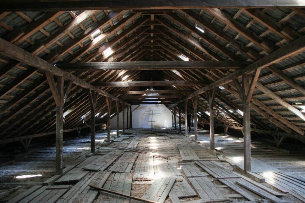 A Detailed Image Of An Old, Abandoned Attic With Wooden Beams Supporting A Slanted Roof. The Floor Is Covered With Scattered Wooden Planks And Dust, With Sunlight Filtering Through Gaps In The Roof. The Background Features A White Brick Wall With A Cross-Shaped Wooden Structure.