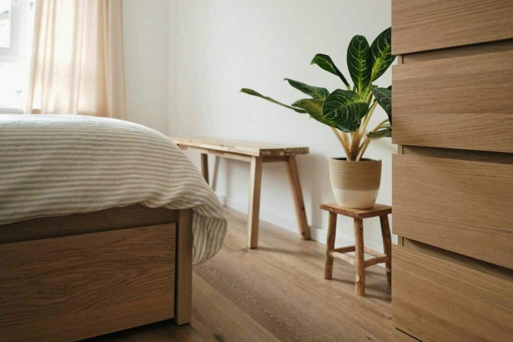 A Detailed Image Of A Minimalist Bedroom With Wooden Furniture, Including A Bed Frame, Dresser, And Bench. A Potted Green Plant Sits On A Small Wooden Stool Beside A White Wall, With Soft Natural Light Coming Through A Window With Beige Curtains.