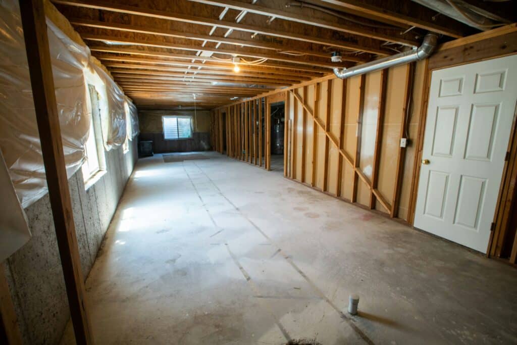 A Wide-Angle View Of An Unfinished Basement With Exposed Wooden Framing, Concrete Floors, Visible Insulation, And Utility Lines. The Space Is Lit By Natural Light From Small Windows And A Single Ceiling Bulb. A White Door Is Installed On One Wall Near A Partially Built-Out Area.