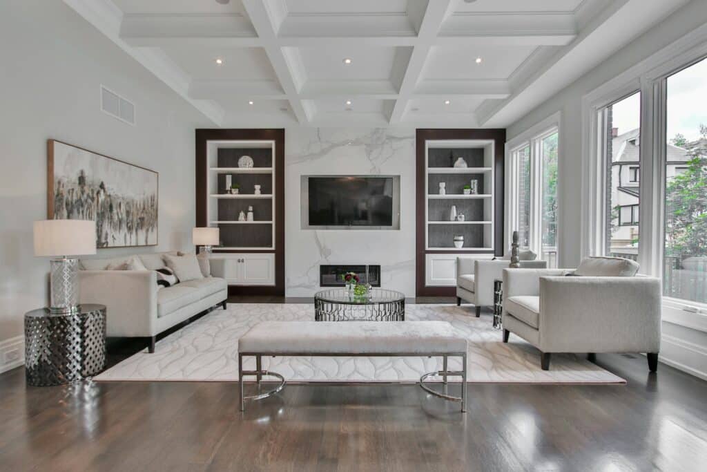 A Bright, Modern Living Room Featuring Neutral-Toned Furniture, A Large Wall-Mounted Tv Centered Between Built-In Shelving Units, And Floor-To-Ceiling Windows That Let In Natural Light. The Space Includes A Patterned Area Rug, A Round Coffee Table With Flowers, And A Coffered Ceiling With Recessed Lighting.