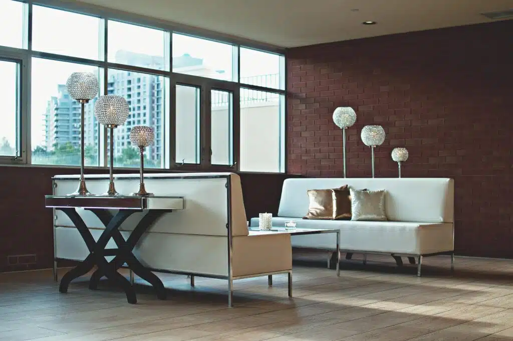 A Modern Apartment Lounge With White Sofas, Decorative Crystal Floor Lamps, And A Mirrored Side Table, Set Against A Brick Accent Wall With Large Windows Overlooking Tall Buildings.
