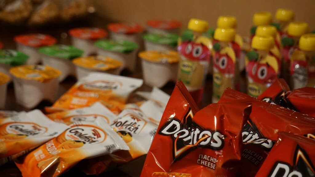 A Close-Up View Of Assorted Packaged Snacks On A Table, Including Bags Of Doritos, Goldfish Crackers, And Various Pudding Cups And Fruit-Flavored Squeeze Pouches, All Arranged In Rows.