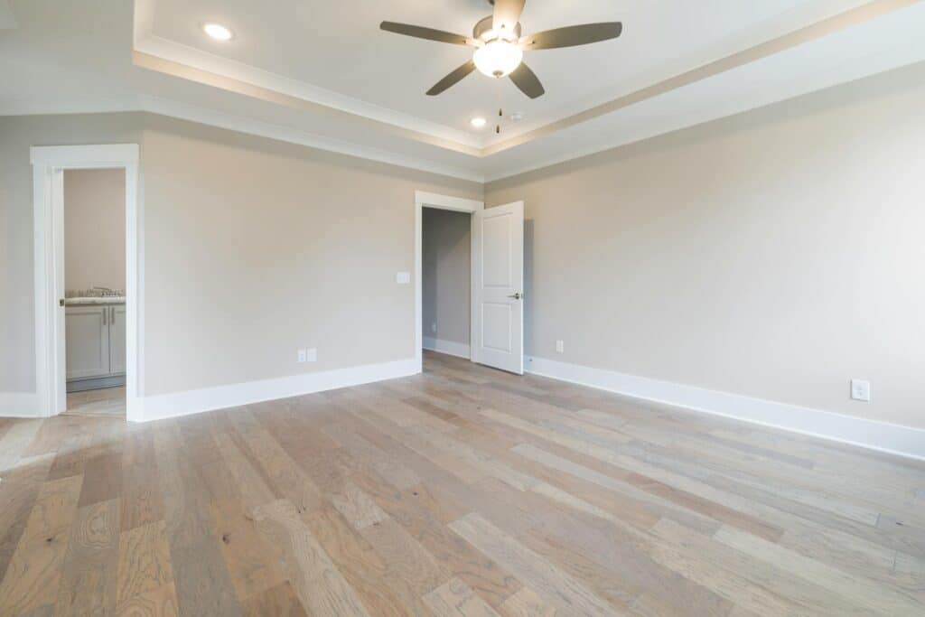 A Spacious, Empty Room With Light Wood Flooring And Beige Walls, Featuring A White-Trimmed Doorway, An Open Door Leading To Another Room, And A Ceiling Fan With Light In The Center Of The Tray Ceiling. Recessed Lighting Highlights The Clean, Modern Aesthetic.