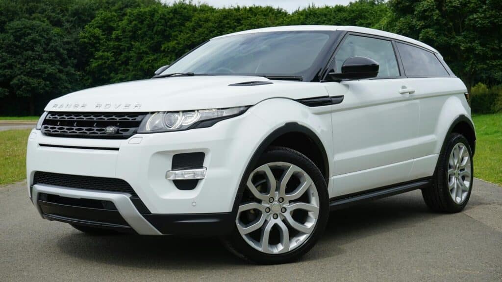 A White Range Rover Evoque Suv Parked On A Paved Road Next To A Grassy Area, With Dense Green Trees In The Background.