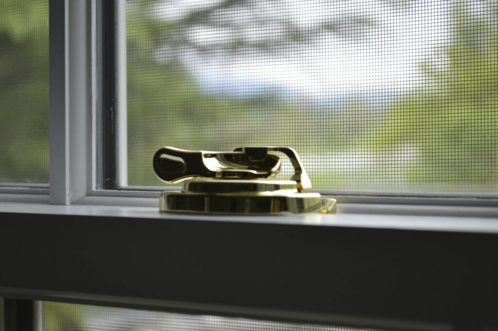 A Close-Up Image Of A Gold Window Lock On A White Window Frame, With A Mesh Screen And A Blurred View Of Green Trees In The Background.