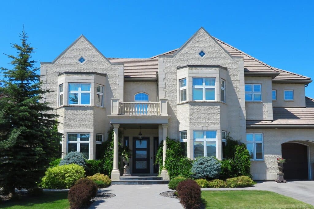 A Front-Facing Image Of A Large Beige Two-Story Brick House With Symmetrical Bay Windows, A Central Balcony Above The Entrance, And Well-Maintained Landscaping Including Shrubs And A Tree Under A Clear Blue Sky.