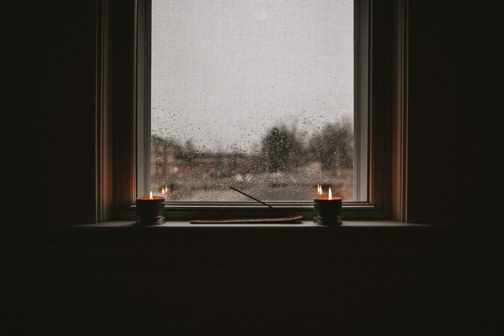 A Dimly Lit Indoor Scene Showing A Rainy Window With Water Droplets On The Glass. Two Lit Candles Sit On The Windowsill, One On Each Side, With A Wooden Incense Holder And An Unlit Incense Stick Placed Between Them. The View Outside Is Blurry, Showing Overcast Skies And Faint Outlines Of Trees And Buildings.