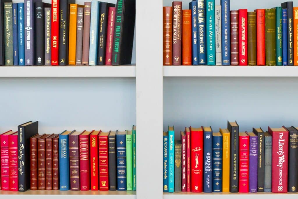 A White Bookshelf Filled With A Vibrant Assortment Of Hardcover Books Arranged Neatly By Color, With Titles In Various Fonts And Sizes Creating A Visually Striking Rainbow Pattern.