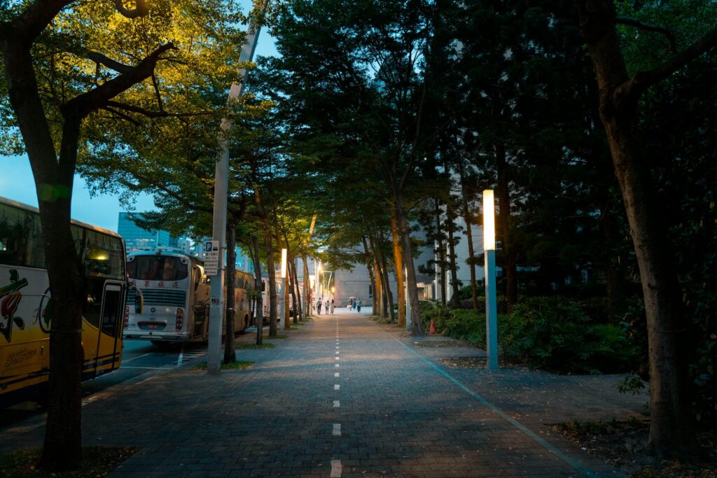 A Paved, Tree-Lined Urban Walkway At Dusk With Soft Street Lighting, Tour Buses Parked On The Left Side Of The Road, And Pedestrians Visible In The Distance Under The Warm Glow Of City Lamps. The Path Is Divided With A Painted White Line, Suggesting Shared Use For Walking And Biking. The Scene Blends Urban Infrastructure With Greenery.