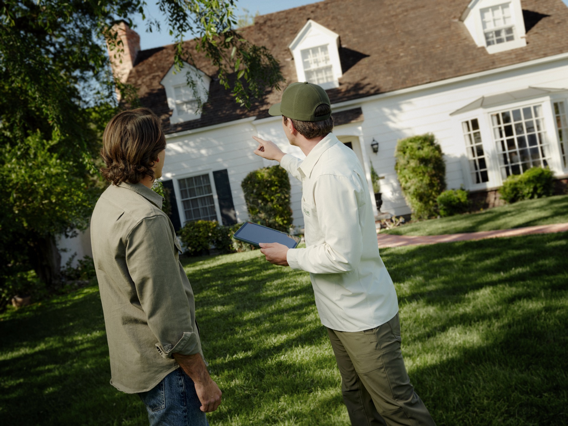 Aptive-Environmental-Specialist-Inspecting-Home-Exterior Aptive Environmental Specialist Holding A Tablet And Pointing Toward A Home'S Exterior While Inspecting The Property With A Homeowner.